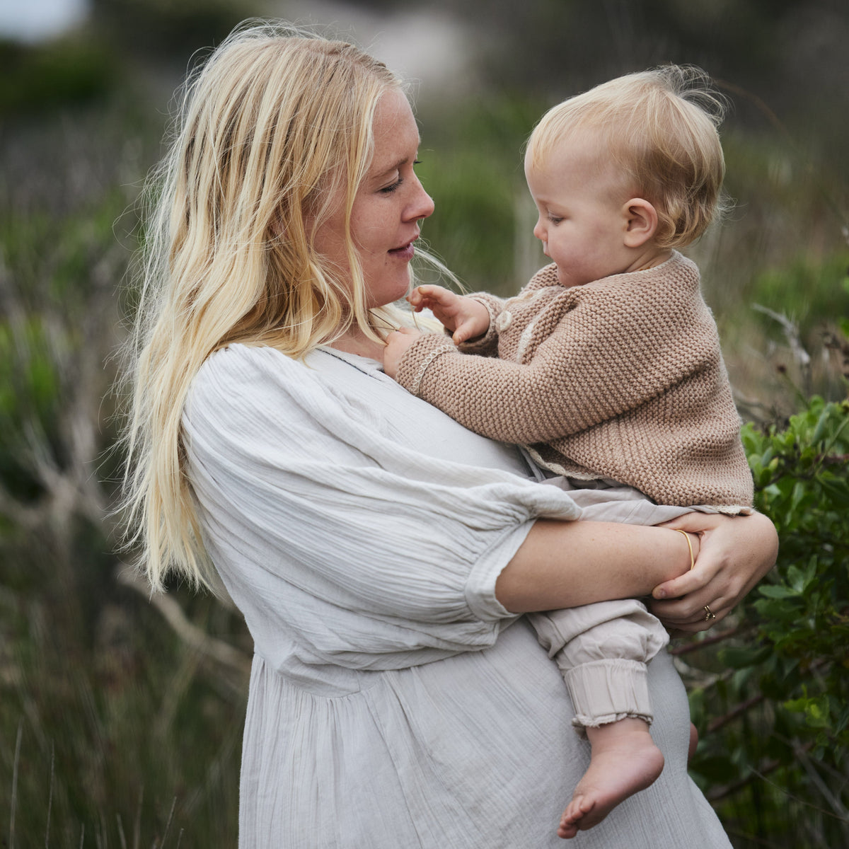 A woman in a pale blue organic cotton dress holds a baby wearing a beige knit sweater and matching pants in an outdoor setting with blurred greenery.