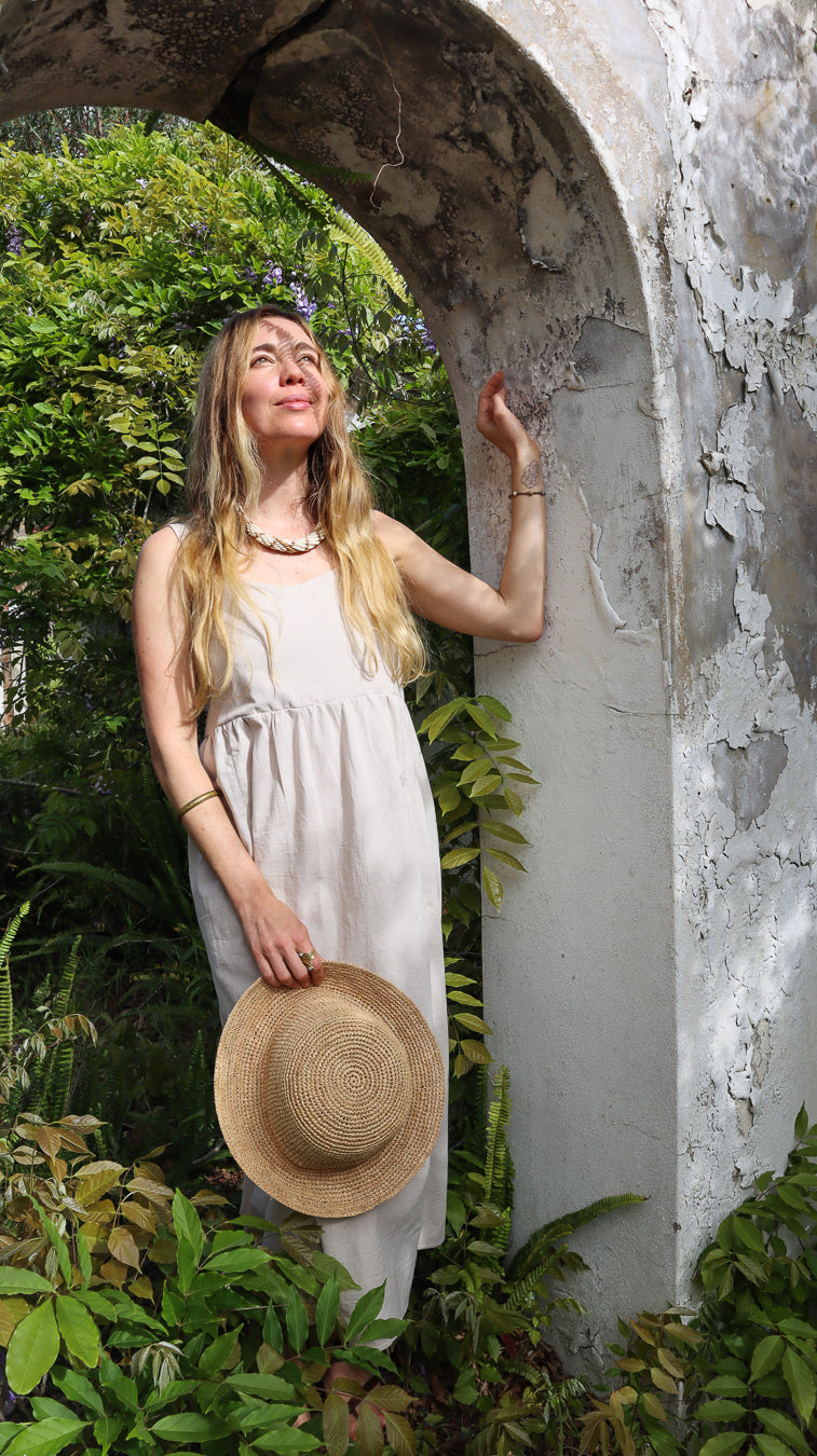 A woman with long blonde hair stands under a weathered stone archway in a garden, wearing a light beige sleeveless dress and holding a straw hat.